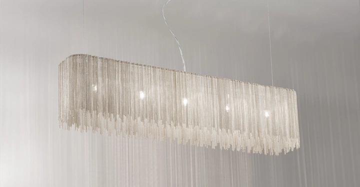 Ein länglicher, rechteckiger Kronleuchter mit Ketten hängt von der Decke. Die hellen Lichter sind von glänzenden, dünnen Metallketten umgeben. Der Hintergrund ist eine graue, glatte Wand.