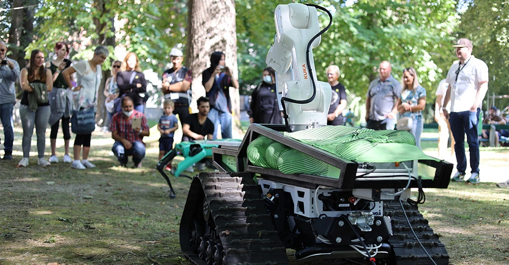 Roboter mit weißem Arm bewegt sich auf Raupenantrieb im Park umgeben von Menschen die zuschauen. Der weiße Arm trägt den Schriftzug KUKA.