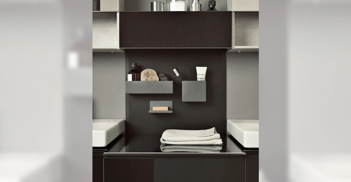 Galerie-Bild 4: Ein Badezimmerregal hält Flaschen Zahnbürsten und Handtücher; es befindet sich zwischen zwei Waschbecken in einem modernen Badezimmer mit grauen Wänden und schwarzen Schränken. Text auf Tube: Aesop Mandarin Facial Hydrating Cream