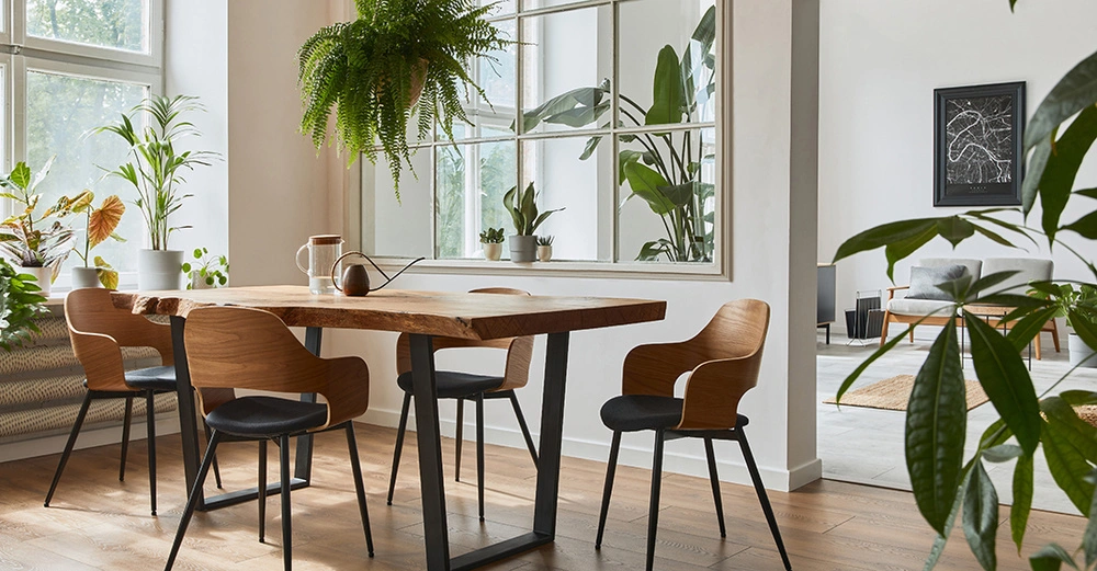 Holztisch mit vier Stühlen stehend in einem hellen Zimmer Pflanzen umgeben den Tisch und draußen durch Fenster ist Grün zu sehen. Im Hintergrund ist ein Wohnzimmer zu erkennen.