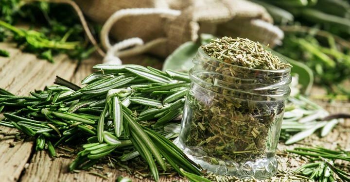 Galerie-Bild 7: Ein Glas mit getrockneten Kräutern steht auf einem hölzernen Tisch. Umgeben von frischen Rosmarinzweigen und einem leeren, umgekippten Jutesack im Hintergrund.