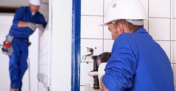 Arbeiter montiert Elektroinstallation in eine verfließte Wand ein zweiter Arbeiter steht auf einer Leiter im Hintergrund beide tragen blaue Overalls und weiße Schutzhelme im Innenraum.