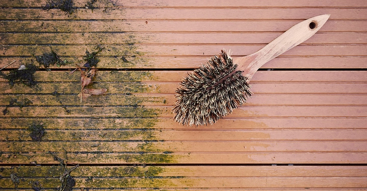 Eine Bürste liegt auf einer Holzterrasse. Die Bürste liegt still. Der linke Teil der Terrasse ist verschmutzt und moosbewachsen während der rechte Teil sauber ist.