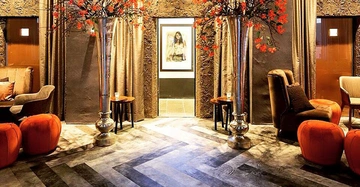 Große silberne Vasen mit roten Blumen stehen in einer gemütlichen Sitzecke in einem Luxushotel mit bequemen Sesseln, Hockern, braunen Vorhängen und einem gerahmten Bild an der Wand.