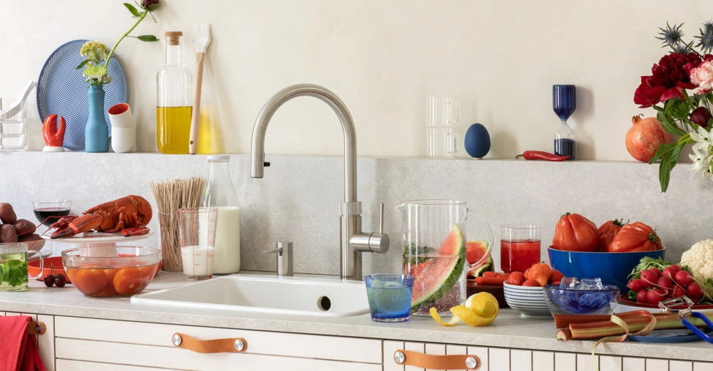 Ein Küchenspülbecken mit Wasserhahn ist umgeben von verschiedenen Lebensmitteln und Küchenutensilien wie Tomaten, Milch, einer Hummer, Obst und Gemüse in einer modernen Küche.