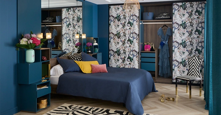 Bett ruht in einem elegant dekorierten Schlafzimmer mit blauem Bettbezug; der Kleiderschrank im Hintergrund hat offene Schiebetüren mit Blumenmustern, die Kleidung und Accessoires zeigen; links steht eine Vase mit Blumen auf einem Regal