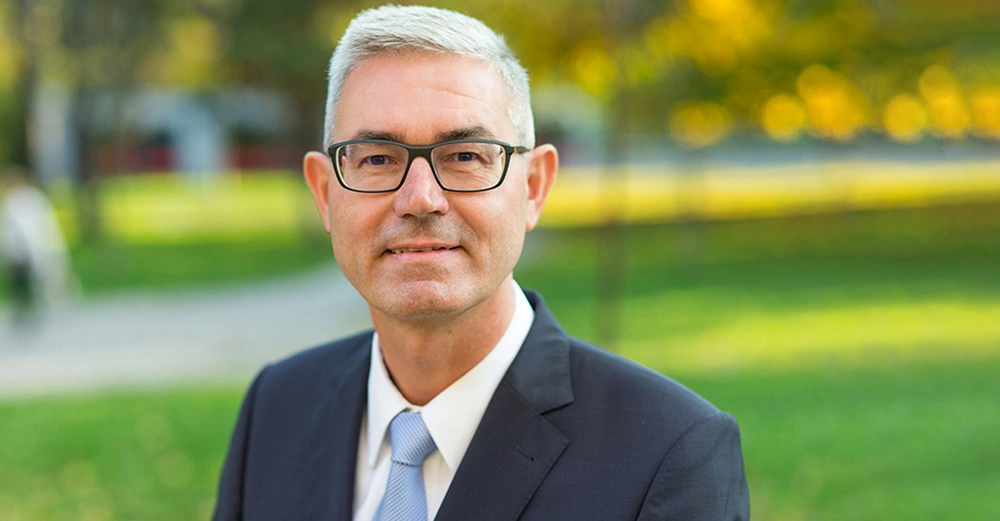 Ein Mann mit grauen Haaren und Brille lächelt leicht in die Kamera im Freien vor einem verschwommenen grünen Hintergrund mit Bäumen und einem Gehweg