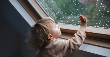 kleines blondes, etwa 2jähriges Mädchen steht unter Dachfenster und berührt die vom Regen nasse Glasscheibe des Fensters. Draußen starker Regen, grau in graue Natur.