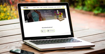 Laptop zeigt eine Webseite mit dem Titel ISOVER SAINT-GOBAIN und Texten SO WIRD GEDÄMMT Für jede Anwendung die passende Lösung und Was möchten Sie dämmen auf Holztisch im Freien