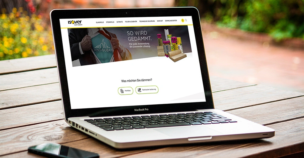 Laptop zeigt eine Webseite mit dem Titel ISOVER SAINT-GOBAIN und Texten SO WIRD GEDÄMMT Für jede Anwendung die passende Lösung und Was möchten Sie dämmen auf Holztisch im Freien