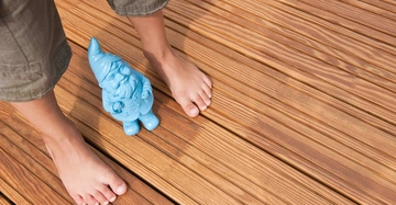 Ein blauer Gartenzwerg steht zwischen zwei bloßen Füßen auf einem hölzernen Terrassenboden.
