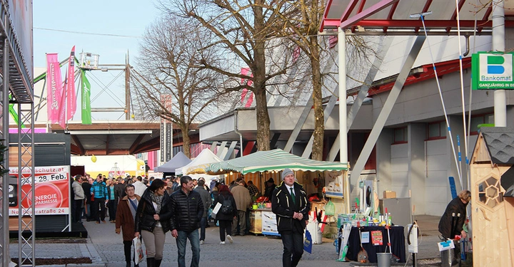 Galerie-Bild 12: Menschen gehen durch eine belebte Outdoor-Messe mit Ständen und Pavillons; einige Bäume und Gebäude sind im Hintergrund sichtbar. Texte in Bild: Schutz.at Energie Sparmesse Bankomat - Ganzjährig in Betrieb.