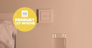 Schalter mit der Aufschrift Deckenleuchte an der Wand über einem Glas Wasser und einem Bett mit der Markierung Produkt der Woche 48 in einem Schlafzimmer.