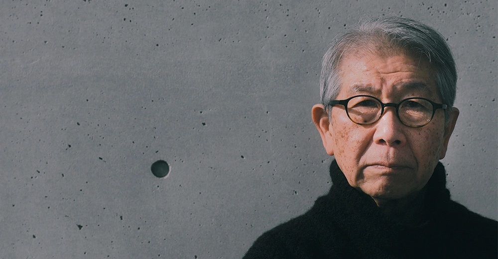 Portrait von Riken Yamamoto, nicht lächelnd, schwarze Hornbrille, schwarzer Rollkragenpulli, im Hintergrund eine blanke Sichtbetonwand. 