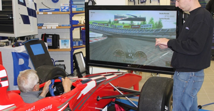 Galerie-Bild 8: Ein Mann sitzt in einem Rennsimulator und fährt ein virtuelles Autorennen auf einem großen Bildschirm während ein anderer Mann daneben steht. Im Hintergrund befinden sich Werkzeuge und Regale. Text: Joe Kranzl Projekt for racing Super Rank Mario Ranola best 1.32.209