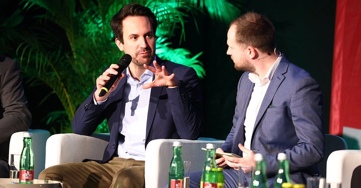 2 Männer in Businessoutfit sitzen mit Mikrofon auf Bühne in weißen Armsesseln und diskutieren.