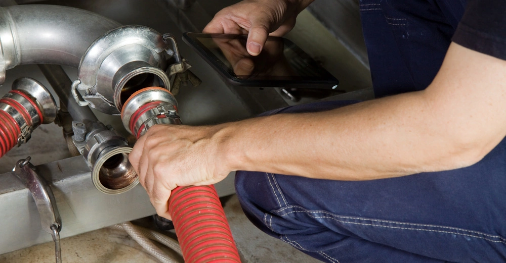 Ein Mensch in Arbeitskleidung hält eine rote Schlauchverbindung und benutzt ein Tablet in einer technischen Umgebung mit Metallrohren und Armaturen.