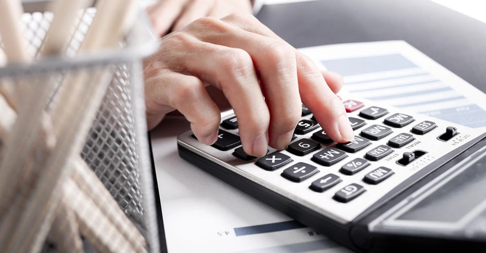 Frau rechnet auf Taschenrechner, zahlreiche Unterlagen und Rechnungen auf Tisch verteilt