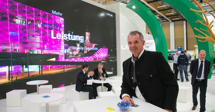 Galerie-Bild 6: Mann lacht und lehnt sich gegen Tisch Schüssel mit Süßigkeiten Messer und Gabel hinten Menschen in Anzügen weißer Stand lila beleuchtetes Gebäude Text auf Leinwand "Leistung" "die Gemeinde"