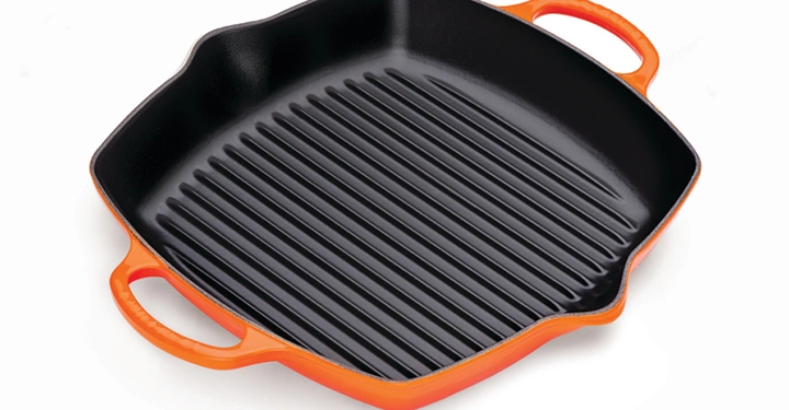 Rote quadratische Grillpfanne mit zwei Griffen liegt auf weißem Hintergrund. Die schwarze Innenseite hat geriffelte Streifen.