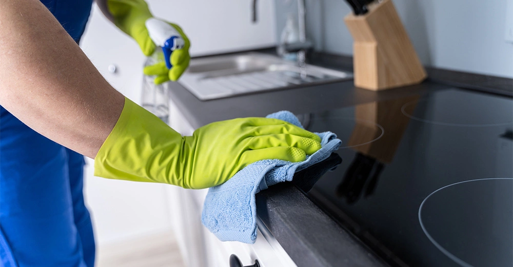 Person mit gelben Handschuhen reinigt eine schwarze Küchenarbeitsfläche mit einem blauen Tuch und einem Reinigungsmittelspender in einer modernen Küche.
