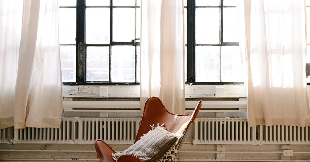 Ein brauner Stuhl steht vor großen Fenstern mit weißen Vorhängen in einem hellen Raum mit Holzboden