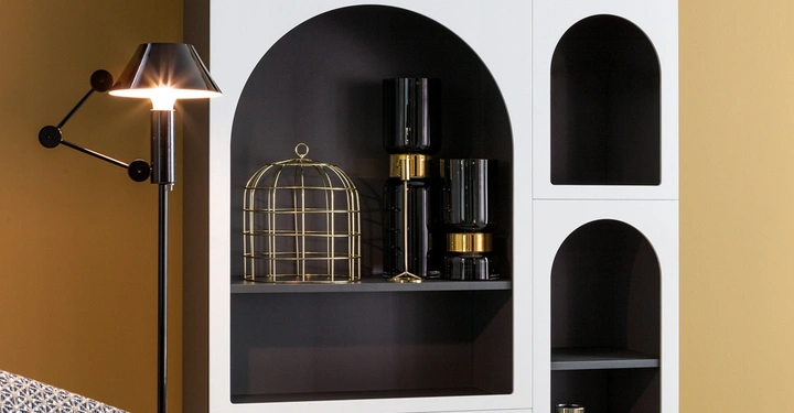 Schwarze Stehlampe leuchtet neben weißem Regal mit abgerundeten Fächern mit Dekorationsobjekten wie goldenen Vogelkasten und schwarzen Vasen in modernem Raum mit gelber Wand.