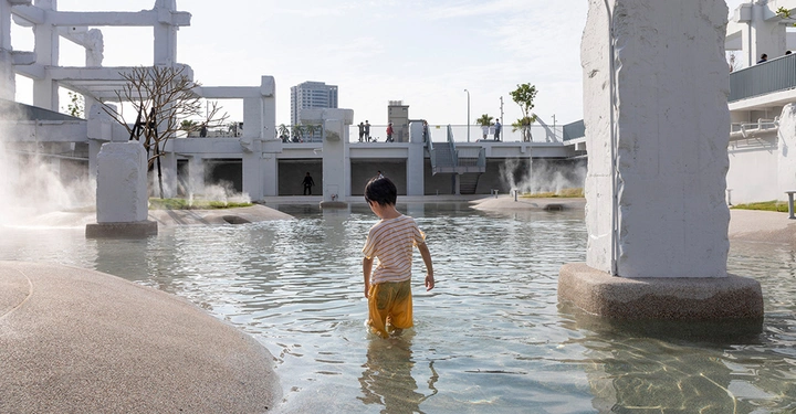 Galerie-Bild 1: Ein Kind watet im knietiefen Wasser eines modernen Wasserparks, umgeben von großen weißen Betonstrukturen und Pflanzen, mit einem Gebäude und mehreren Personen im Hintergrund