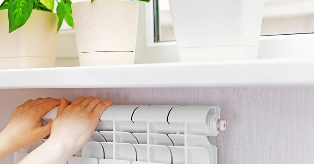 Hände berühren einen weißen Heizkörper in einem Raum mit blassen Wänden Pflanzen in weißen Töpfen stehen auf der Fensterbank oberhalb des Heizkörpers neben einer Fensterscheibe