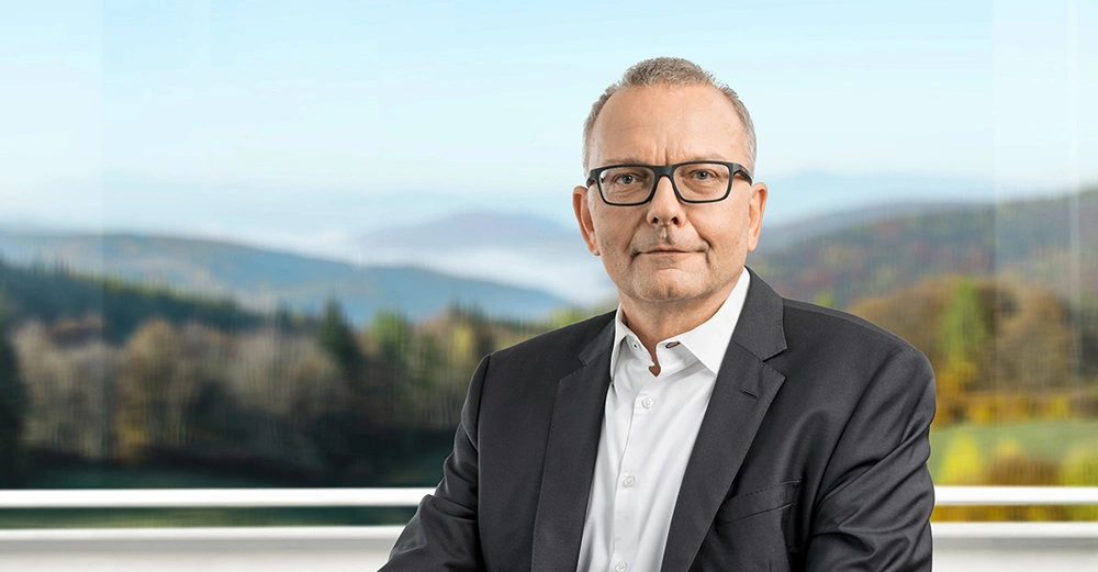 Mann trägt Brille und Anzug und sitzt vor einem Fenster mit nebeligen Hügeln und Bäumen.