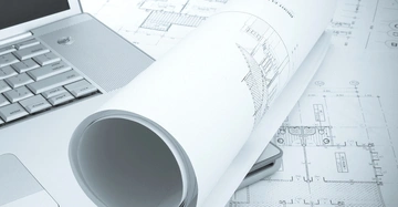 Laptop liegt teilweise geöffnet neben gerollten Architekturzeichnungen auf großformatigen Bauplänen. Designzeichnungen zeigen Gebäudedetails und Maße.