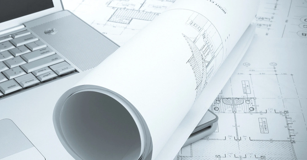 Laptop liegt teilweise geöffnet neben gerollten Architekturzeichnungen auf großformatigen Bauplänen. Designzeichnungen zeigen Gebäudedetails und Maße.