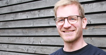 Mann lacht vor einer Holzverkleidung trägt eine Brille und hat kurze blonde Haare quer gestreiftes Holz im Hintergrund