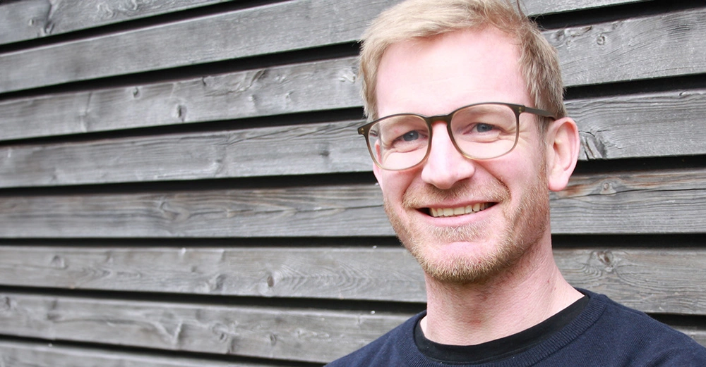 Mann lacht vor einer Holzverkleidung trägt eine Brille und hat kurze blonde Haare quer gestreiftes Holz im Hintergrund