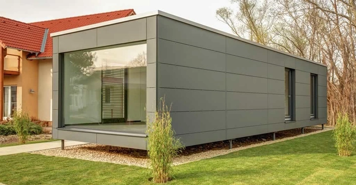 Ein graues Containerhaus steht auf gestütztem Fundament, umgeben von gepflegtem Rasen und Pflanzen, in der Nähe eines Gebäudes mit rotem Dach und Bäumen im Hintergrund.