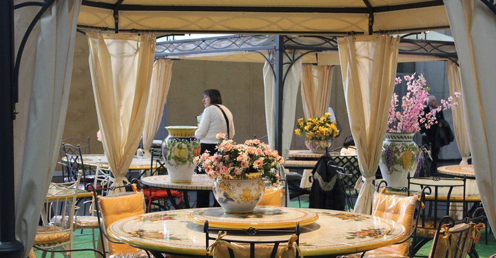 Galerie-Bild 7: Großer Blumentopf mit Blumen steht auf einem dekorativen Tisch Menschen sind umgeben von Pavillons mit cremefarbenen Vorhängen und grüner Teppichboden umarmen einen Innenbereich.