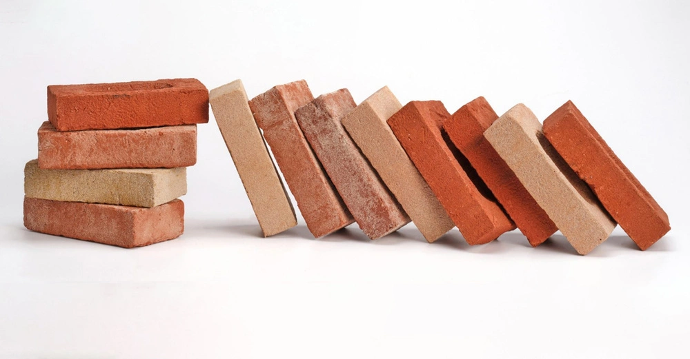 Backsteine liegen in unterschiedlicher Anordnung auf einer weißen Oberfläche Einige sind zu einem Stapel aufgeschichtet und andere lehnen aneinander in einer fallenden Dominoreihe.