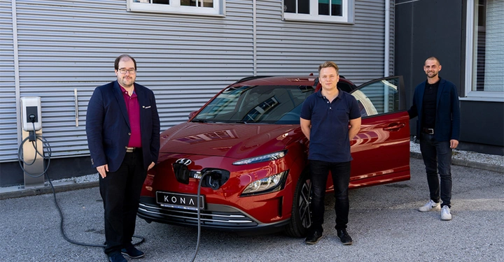 Ein rotes Elektroauto wird von drei Männern in Geschäftskleidung in einem städtischen Bereich gezeigt während es an einer Ladestation aufgeladen wird. Das Nummernschild des Autos zeigt KONA.