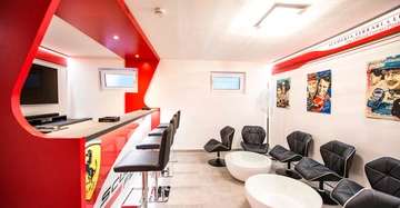 Ein modern gestalteter Raum enthält eine rote Bartheke mit vier schwarzen Hockern zwei kleine Fenster und mehrere Rennfahrerposter an den Wänden Über der Bartheke steht SCUDERIA FERRARI CLUB The Official Ferrari Passione.
