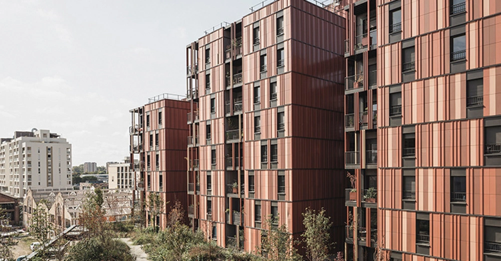 Mehrere hohe Wohngebäude mit rot-orangen Fassaden stehen nebeneinander. Im Hintergrund sind weitere Gebäude sichtbar und im Vordergrund wachsen Bäume und Sträucher. Klarer Himmel.