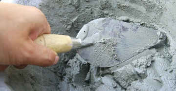 Hand hält Kelle rührt Zementmischung in einem Behälter vorbereitend für Bau oder Renovierungsarbeiten gräuliche Textur sorgt für glatte Verteilung im umgebenden Container.