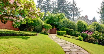 Ein gepflegter Gartenweg aus Steinplatten führt zwischen grünen Büschen und blühenden Sträuchern vorbei an einem Backsteinhaus Richtung Bäume im Hintergrund.