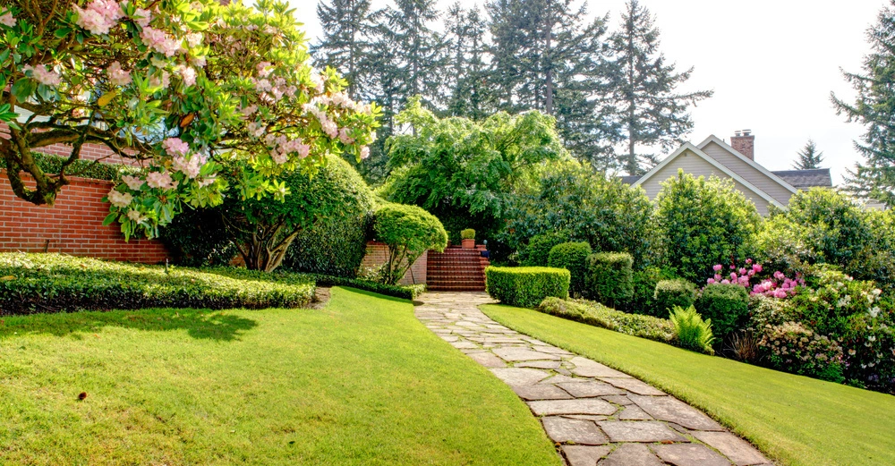 Ein gepflegter Gartenweg aus Steinplatten führt zwischen grünen Büschen und blühenden Sträuchern vorbei an einem Backsteinhaus Richtung Bäume im Hintergrund.