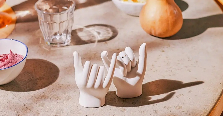 Zwei weiße Hand-Statuen zeigen Gesten auf einem Tisch mit einem Glas einer Schale mit Hummus einer Schale mit Essen und einer Kürbis-Dekoration.