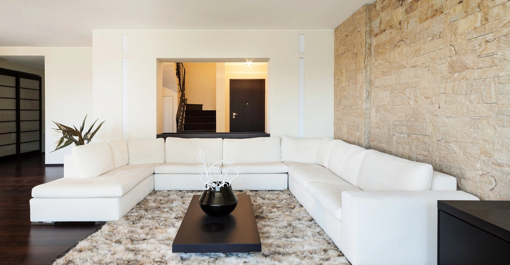 Weißes Ecksofa steht auf einem flauschigen Teppich vor einer Steinwand weiße Vase auf schwarzem Couchtisch Hintergrund zeigt einen offenen Durchgang zu einem Flur mit Treppe und Tür