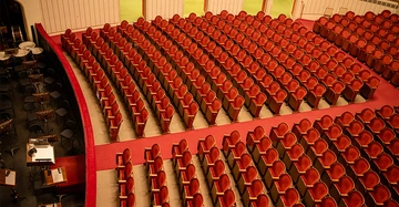 Rote Theatersitze in Reihen angeordnet stehen leer in einem großen Saal mit rotem Teppichboden und einem Orchesterbereich links mit Musikinstrumenten und Stühlen