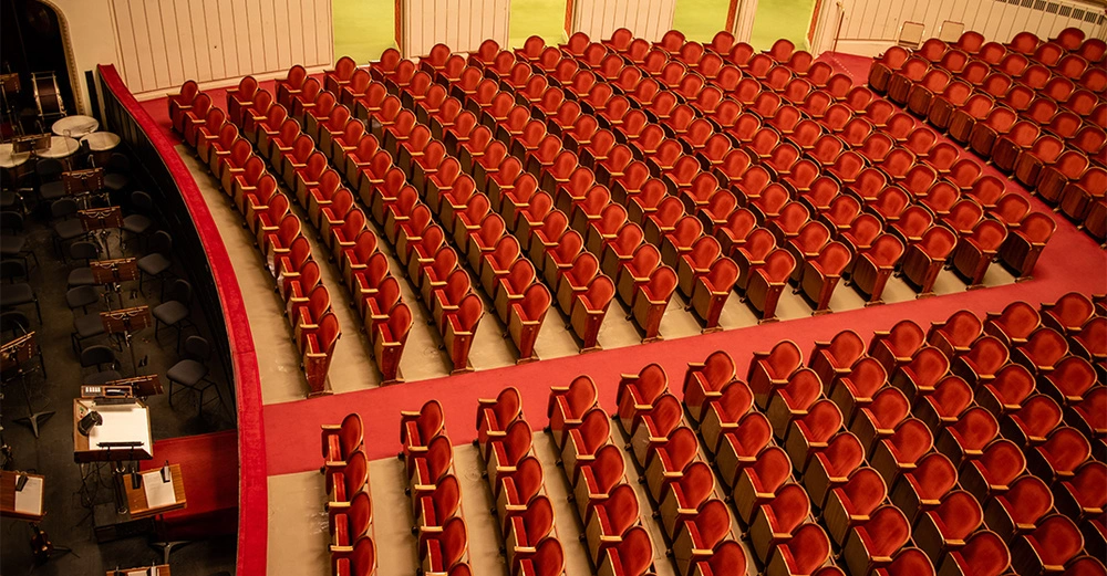 Rote Theatersitze in Reihen angeordnet stehen leer in einem großen Saal mit rotem Teppichboden und einem Orchesterbereich links mit Musikinstrumenten und Stühlen