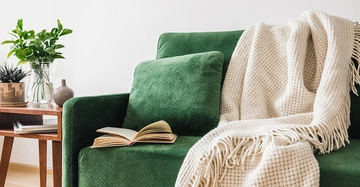 Wohnzimmer mit grünem Sofa, Kissen, Buch und Decke. Holztisch mit Pflanzen sorgt für natürliche, wohnliche Atmosphäre.