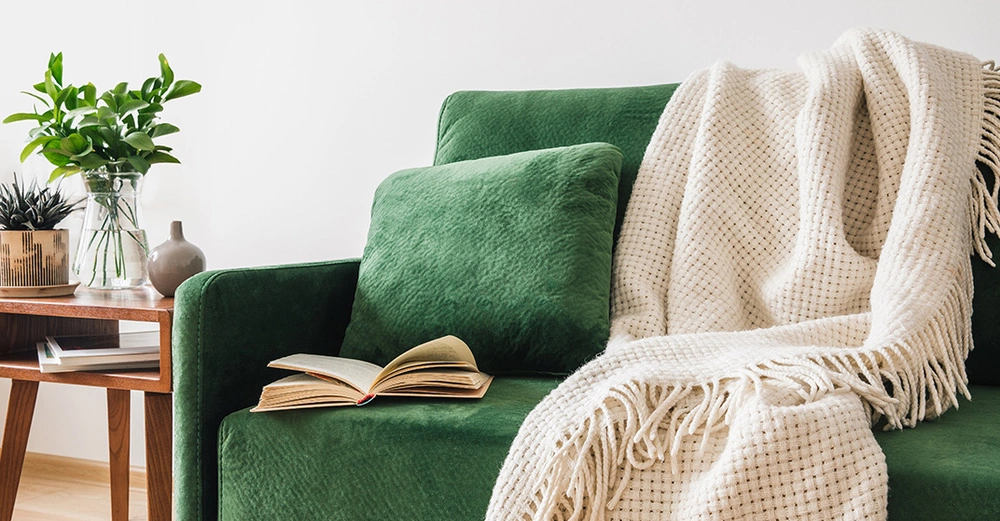 Wohnzimmer mit grünem Sofa, Kissen, Buch und Decke. Holztisch mit Pflanzen sorgt für natürliche, wohnliche Atmosphäre.