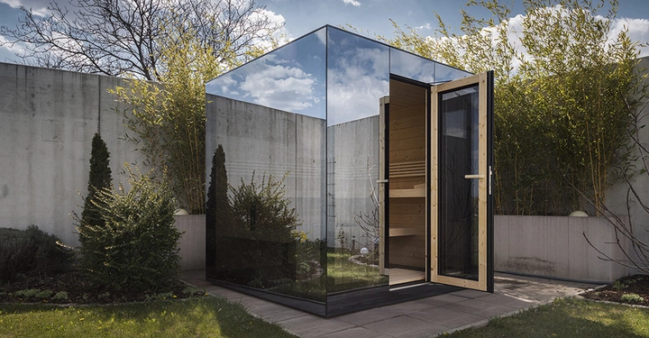 Ein verspiegelter Würfel mit geöffneter Holztür steht in einem Garten umgeben von Betonwänden, Pflanzen und Gras unter einem bewölkten Himmel.